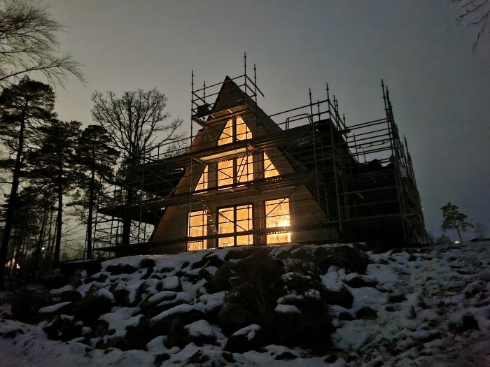 A-formad byggnad på natten med upplysta fönster och byggnadsställningar, omgiven av snö och träd.