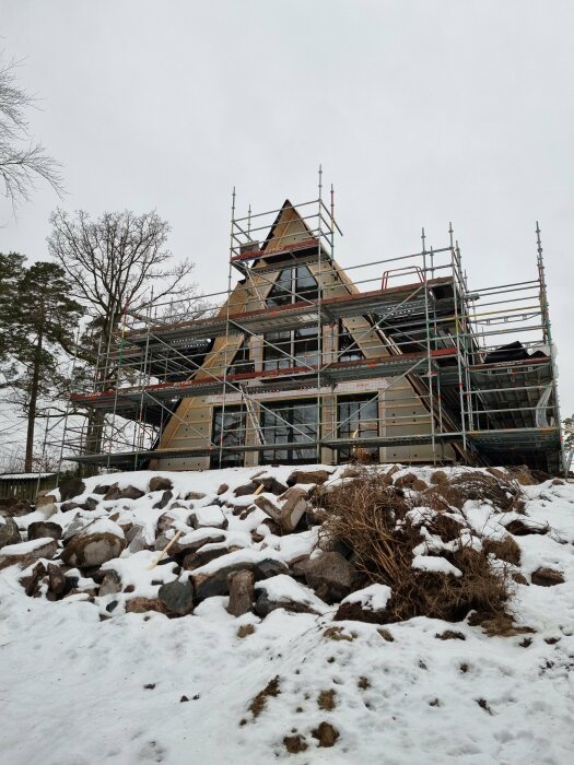 Ett hus under byggnation med ställningar, snötäckta stenar i förgrunden och flerparten av fönstren installerade.