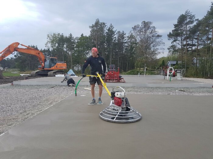 Byggarbetare jämnar ut nylagd betong med en helikopterglättare på en byggarbetsplats. En grävmaskin syns i bakgrunden.