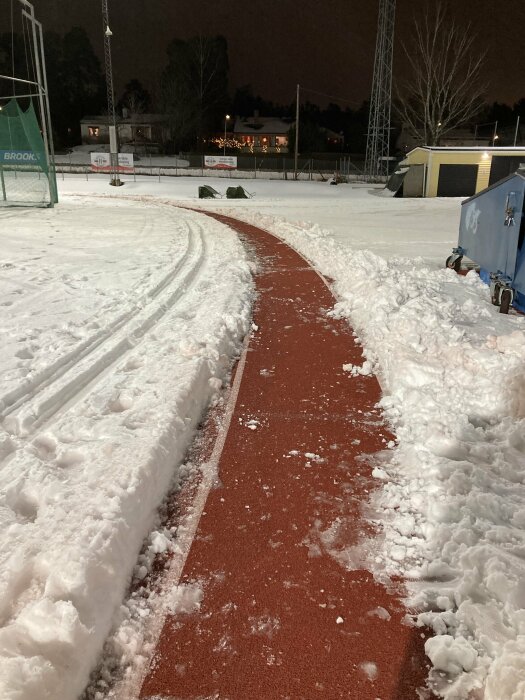 Snötäckt idrottsplats med skottad röd löparbana i centrum, snödrivor på sidorna och byggnader i bakgrunden i kvällsljus.