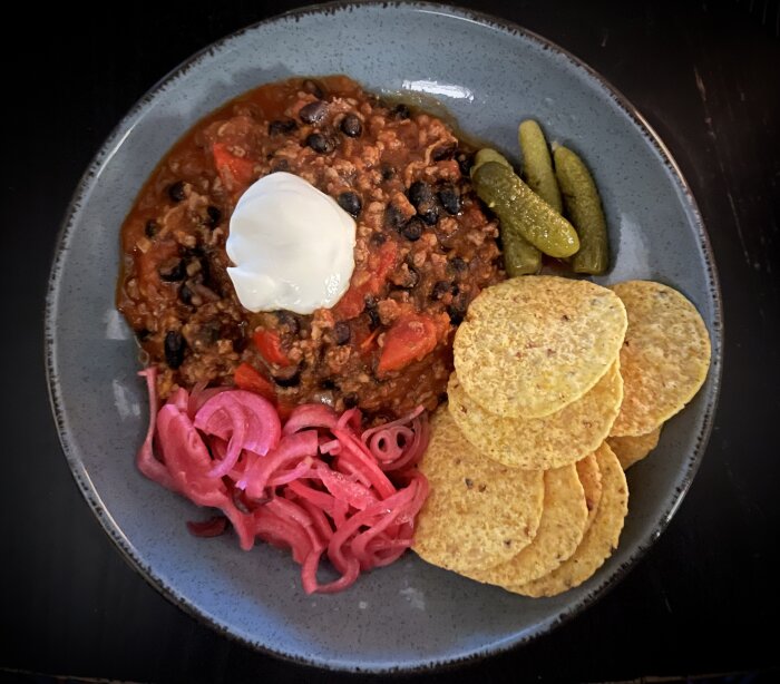Tallrik med chili, gräddfil, picklad rödlök, cornichoner, nachos och en brownie till dessert.