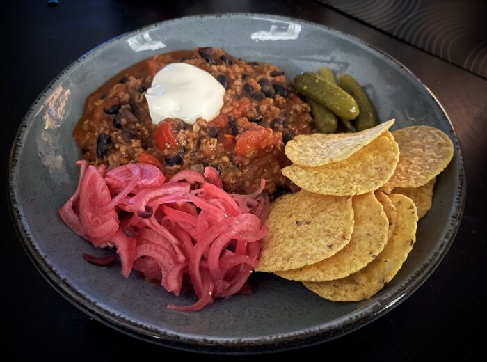 Tallrik med chili toppad med crème fraiche, picklad rödlök, cornichoner och nachos.