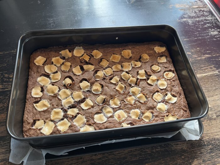 Brownie i en bakform, toppad med smälta marshmallows och inslag av choklad och nötter, stående på ett mörkt träbord.