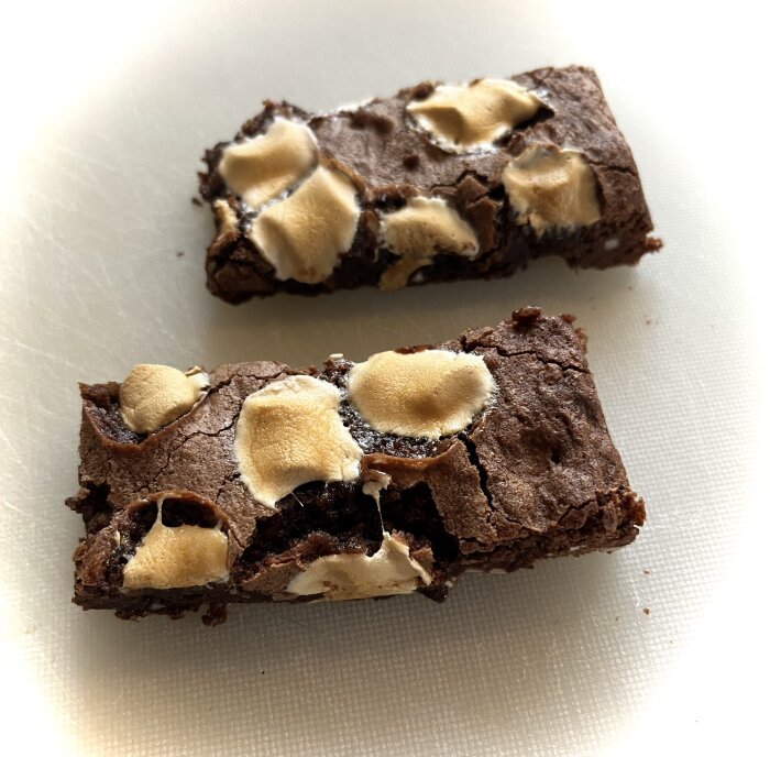 Två bitar brownie med jordnötter, cashewnötter, hackad choklad och minimarshmallows på en skärbräda.