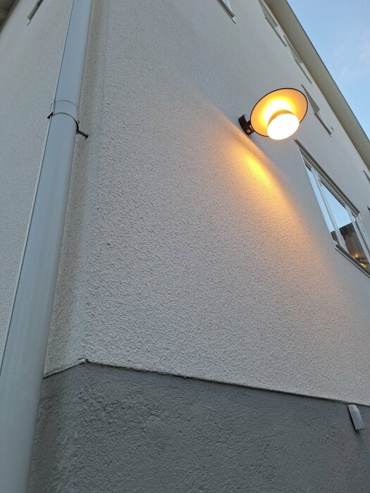 Vit stänkputsad fasad med synlig struktur, en orange vägglampa lyser upp ytan nära ett fönster, fotad nära för detalj.