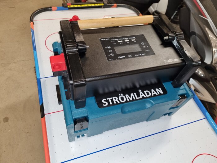 Bärbar strömlåda med svart 2000W inverter ovanpå, placerad på ett bord. Trälaborrat handtag, blå låda märkt "STRÖMLÅDAN".