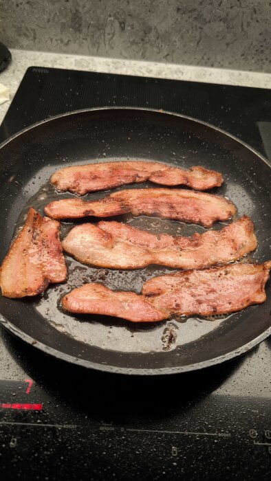 Stekta baconskivor i en svart stekpanna på en spishäll.