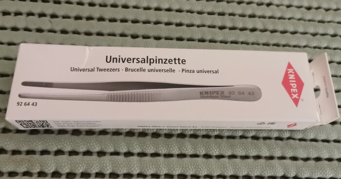 Förpackning med Knipex universalpincett i rostfritt stål, liggande på en grön yta.