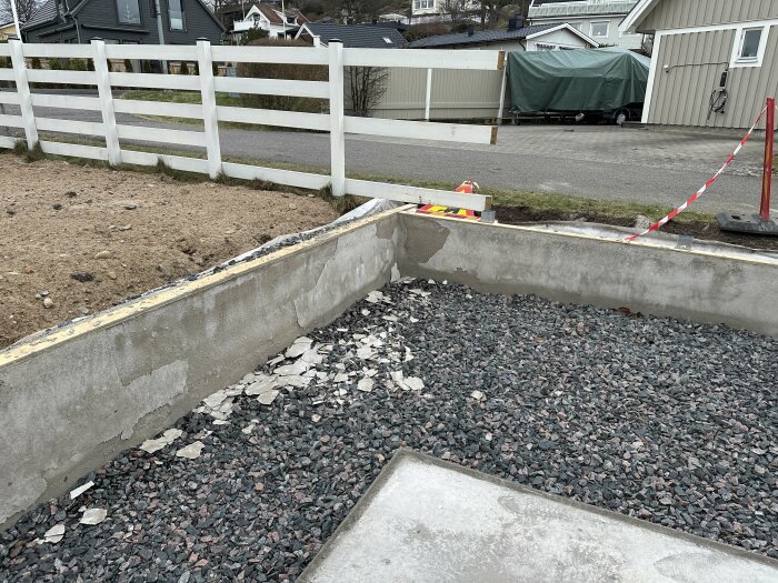 Mur med avflagnad puts, delvis bortblåst efter storm. Grus ligger vid basen, omgiven av staket och byggnader i bakgrunden.