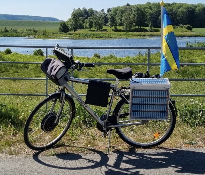 Elcykel med batteripaket och svensk flagga vid sjö och gröna fält i bakgrunden.