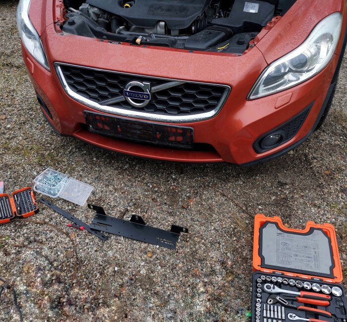 Motorhuven på en röd Volvo öppen, omgiven av verktygslåda och skruvsats på marken, redo för montering eller arbete.