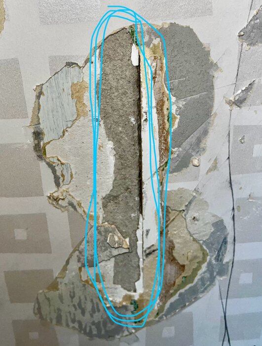 Skadat material på vägg med synliga lager av gammal tapet och möjligen eternit, märkt med blå markering, från 1970-talets husbyggnad.