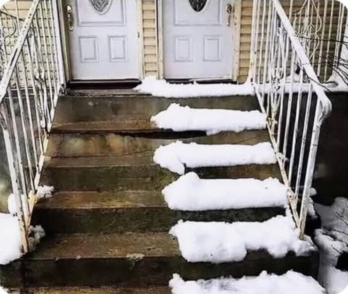 Trappa med snö på varje trappsteg utanför två dörrar, med ett vitt staket på sidorna.