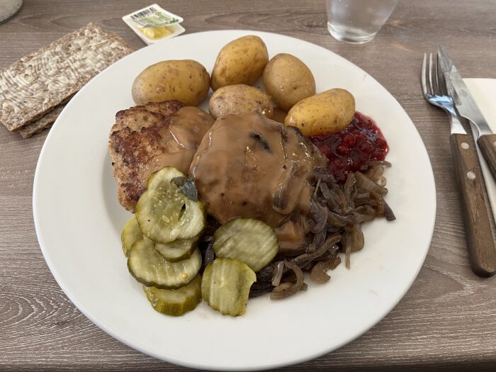 Tallrik med pannbiff, lök, kokt potatis, inlagd gurka och lingonsylt. Hårdmacka bredvid.