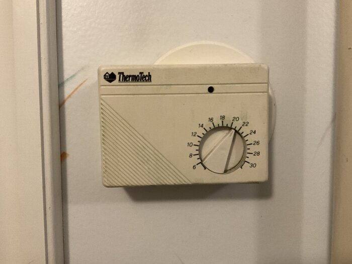 Gammal elektronisk termostat från Thermotech med ratt för temperaturinställning, monterad på en vit vägg.