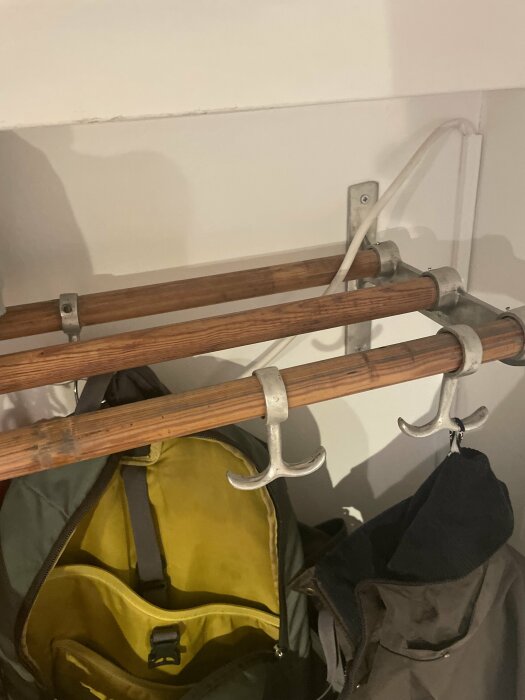 Vägghängd hatthylla i furuträ med metallkrokar och ryggsäckar, visar infästning och träets mönster.