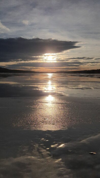 Solnedgång över en frusen sjö med moln som reflekteras på isen, vilket ger ett skimrande ljus över ytan.