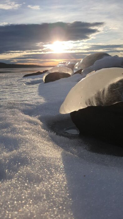 Solnedgång över ett snötäckt landskap med isbeklädda stenar i förgrunden.