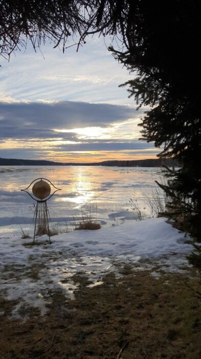 Solnedgång över en frusen sjö med snötäckt strandkant och träd i förgrunden. En dekorativ metallskulptur pryder stranden.