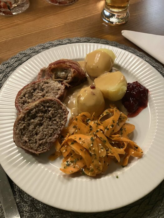 Tallrik med baconlindad köttfärslimpa, potatis med sås, lingon och picklad morot med sesamfrö och koriander. Glas med dryck i bakgrunden.