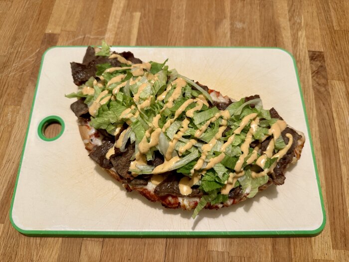 Pizza toppad med kebabkött, sallad och dressing på en skärbräda.