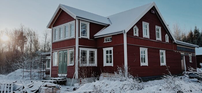 Rött hus med symmetriska vita spröjsade fönster, täckt av snö i vintrigt landskap.