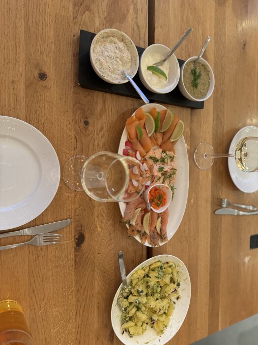 En dukning med lax, lime, såser och potatissallad på ett träbord. Två vinglas och bestick finns också på bordet.