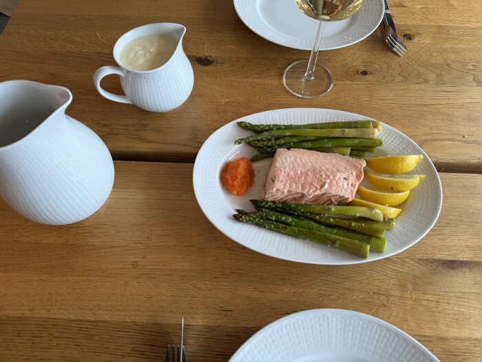 Tallrik med laxfilé, sparris och citronklyftor. Gräddsås i kanna och glas vitt vin på träbord.