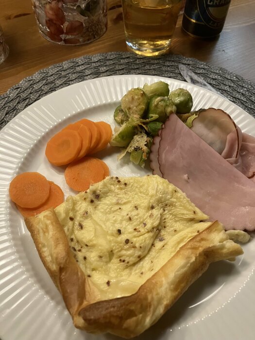 Tallrik med ugnspannkaka, korn av chiliflakes, brysselkål med parmesan, morotskivor och basturökt skinka.