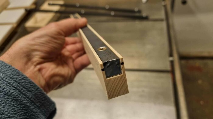 Hand som håller en trälist med infälld stålprofil, vilket ger extra stabilitet och möjlighet att dölja konstruktionens brister.