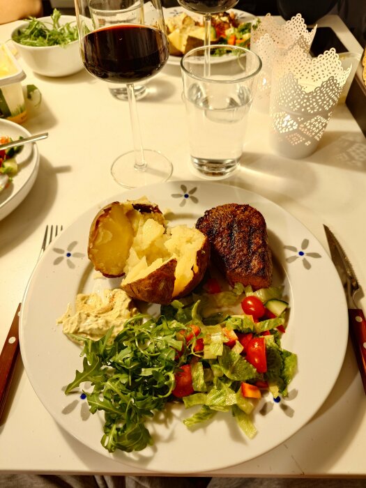 Tallrik med bakad potatis, grillad rostas, sallad och vitlöksröra, serverad med rött vin och vatten.