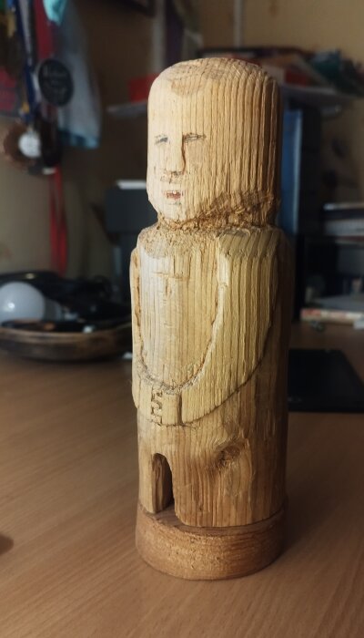 Träfigur skulpterad till en människoliknande form med enkla ansiktsdrag, placerad på ett träbord med en diffus bakgrund inomhus.