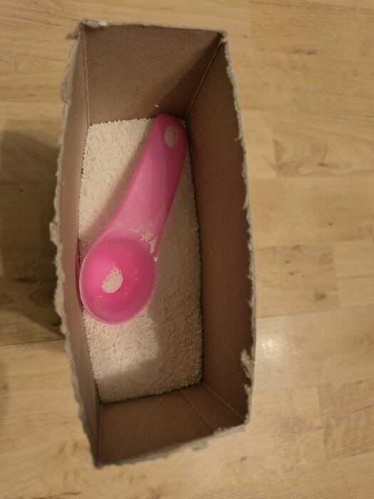 Kartong med diskmedelspulver och en rosa skopa inuti, fotograferad ovanifrån på ett trägolv.