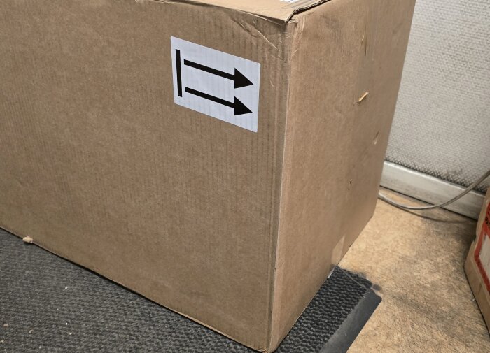 Stor brun kartong med etikett som visar vilka sidor som ska vara uppåt. Kartongen står på en svart matta i ett rum med ljusbrun golvyta.