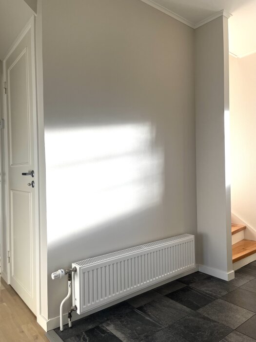 Vit vägg med element i en hall, bredvid en dörr och trappa. Grått klinkergolv, element placerat längs väggen.