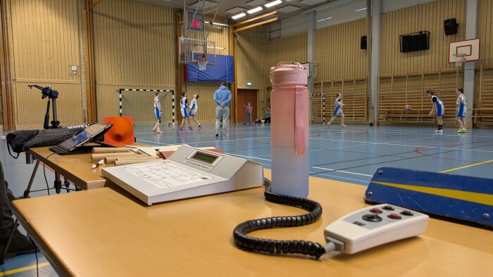 Basketmatch med Söderköping mot Eskilstuna, sett från sekretariatet med utrustning, vattenflaska och spelare på plan i en gympasal.