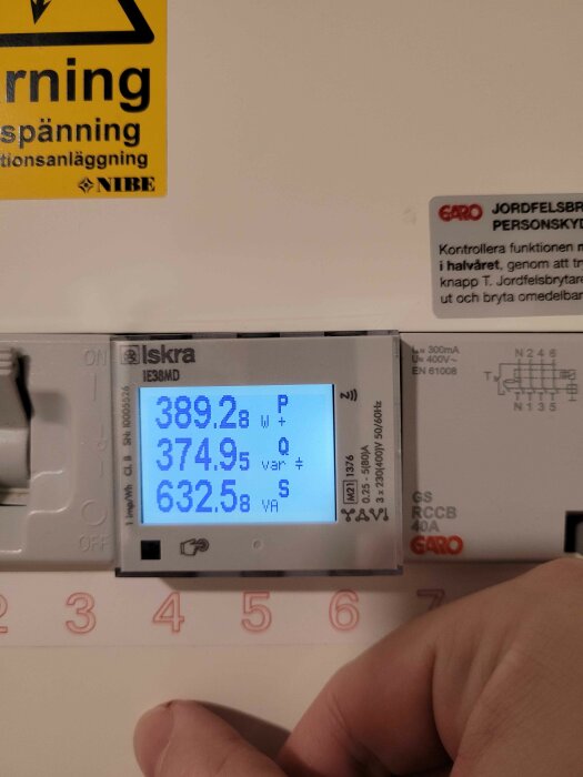 Energimätaren Iskra IE38Mx med avläsningar på skärmen: 389.2 P (W), 374.9 Q (var), 632.5 S (VA). Varningsskylt och instruktion vid sidan.