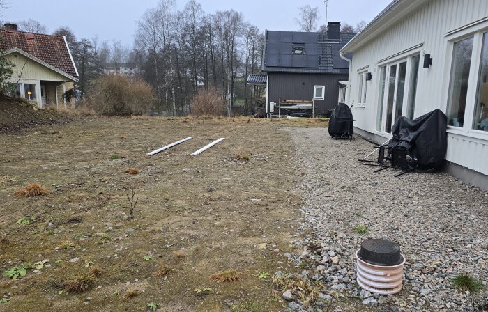 Tomt med grus och markvegetation vid husvägg; två stänger ligger på marken. Huset har stora fönster mot en öppen yta.