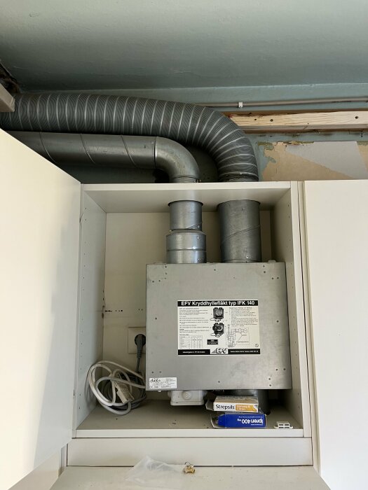 Köksfläkt med anslutna ventilationsrör i ett skåp, märkt "EFV Krydhyllafläkt typ IFK 140", omgiven av kablar och bruksföremål.
