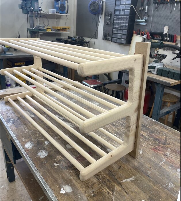 Trähylla med runda stavar, placerad på ett arbetsbord i en verkstadsmiljö, visar steg i ett pågående byggprojekt.