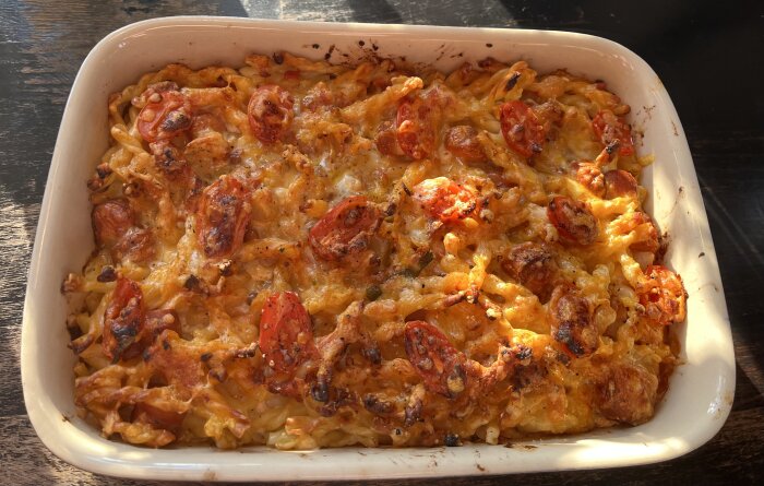 Makaronipudding med tomater och ostgratinering i en vit ugnsform på ett träbord.