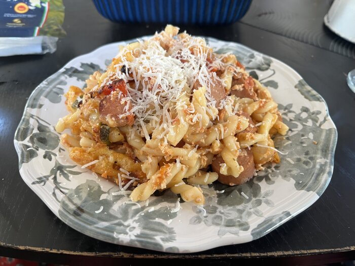 Makaronipudding med korv och tomat, toppad med riven parmesanost, serverad på en dekorativ tallrik.