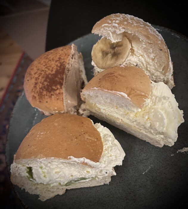 Fyra specialsemlor: en med banan, en med marängswiss, en med pistage och en med citron/lemoncurd, serverade på ett mörkt fat.