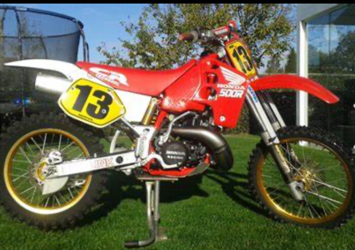 Röd Honda CR 500 motorcykel med nummer 13 på gul bakgrund, placerad på gräs framför träd och ett hus.