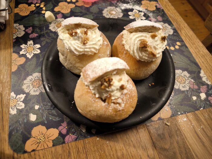 Tre nybakade semlor på en svart tallrik, fyllda med grädde och toppade med mandelkrokant, står på ett blommigt underlägg av trä.