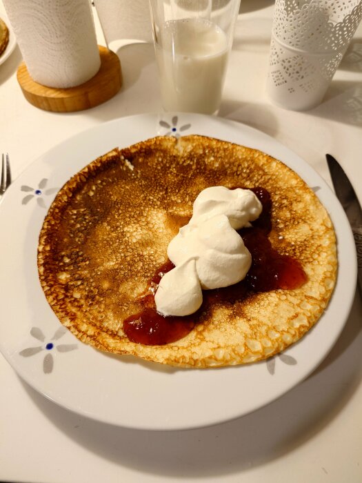 Pannkaka på en tallrik med sylt och mycket grädde, ett glas mjölk i bakgrunden.