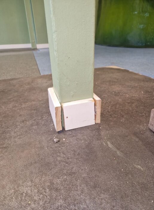 En grön stolpe står på ett mörkt golv, stabiliserad med vita träplattor vid basen.