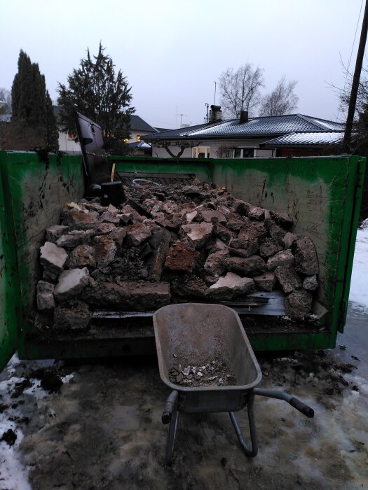 Container fylld med bitar av krossad betong och en skottkärra med kvarvarande betong, omgiven av snötäckta ytor och hus i bakgrunden.