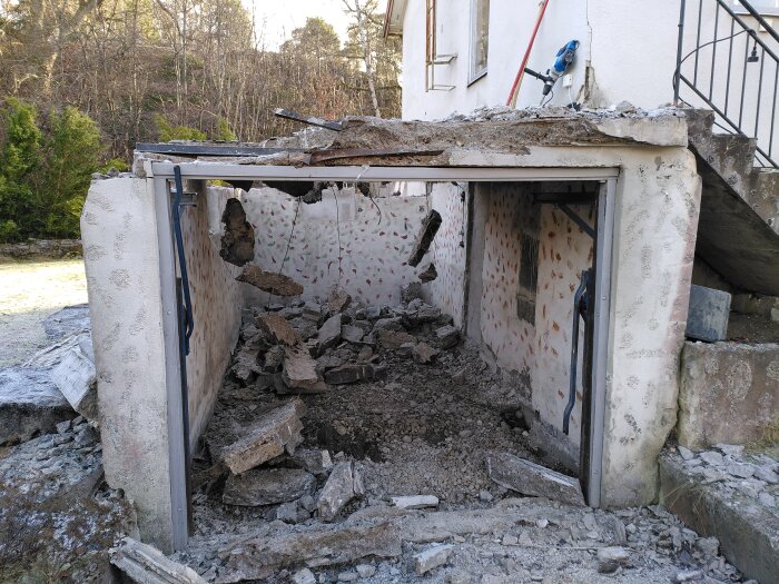 Rivet garage med delvis kollapsat betongtak och synligt grus, rester av väggar och verktyg, omgiven av träd och ett hus med trappa.