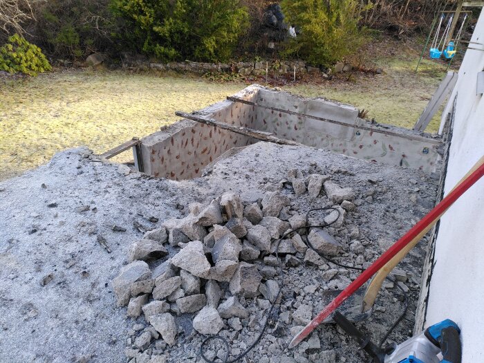 Hög med betongrester från rivet tak och väggar, del av rivningsprojekt med synlig armering och verktyg, trädgård i bakgrunden.